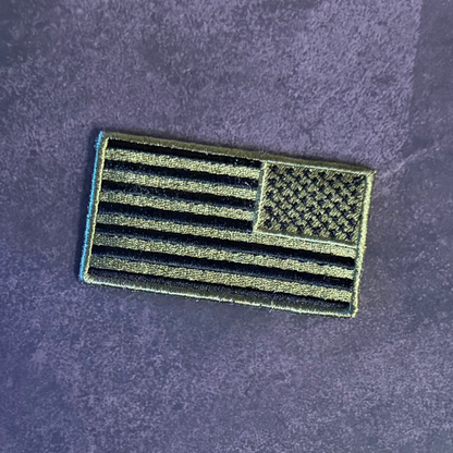 An embroidered American flag patch with black and olive drab stripes and canton, shown on a textured dark purple background.