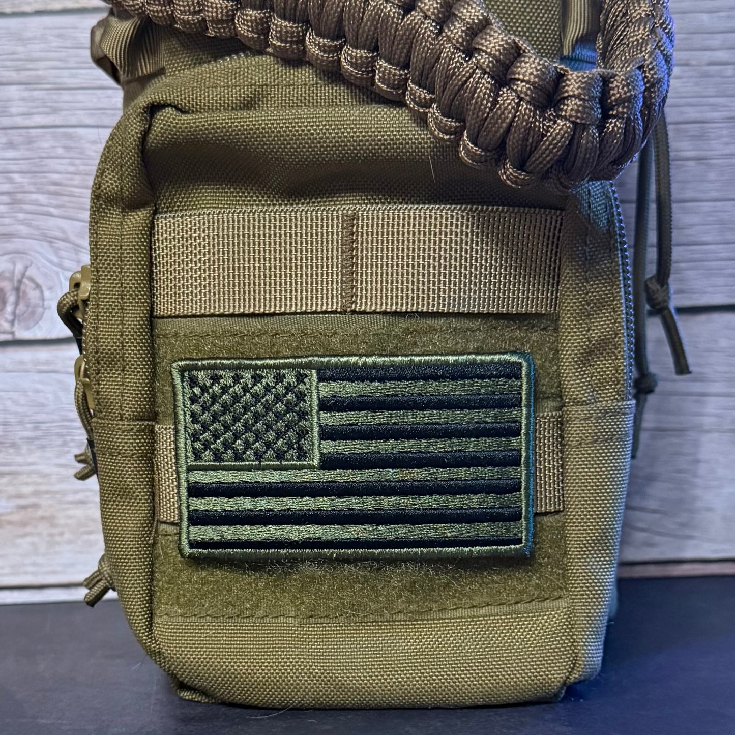An olive drab tactical pouch with a tan webbing strap and a black and green embroidered American flag patch on the front, shown against a wood plank background.