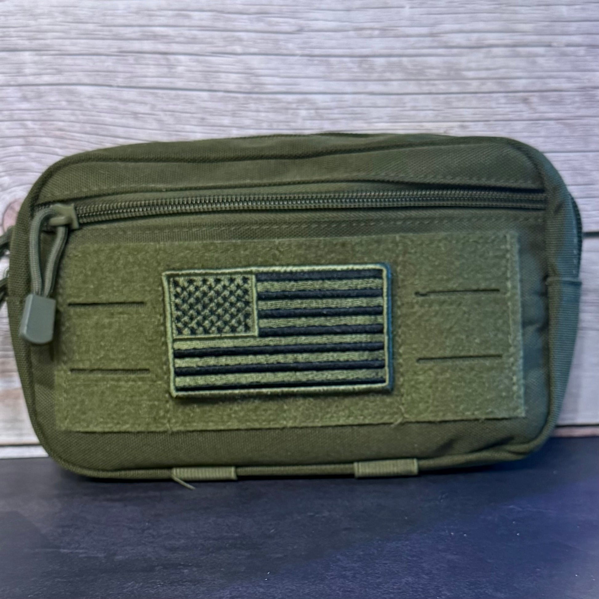 An olive green tactical pouch with a front zippered pocket and a raised American flag patch, shown against a light wood background.