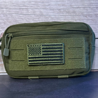 An olive green tactical pouch with a front zippered pocket and a raised American flag patch, shown against a light wood background.