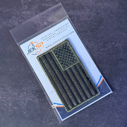 A dark olive green American flag patch with a raised texture, packaged in a clear plastic sleeve with a white backing card featuring the JEKSP brand logo, set against a dark gray textured background.