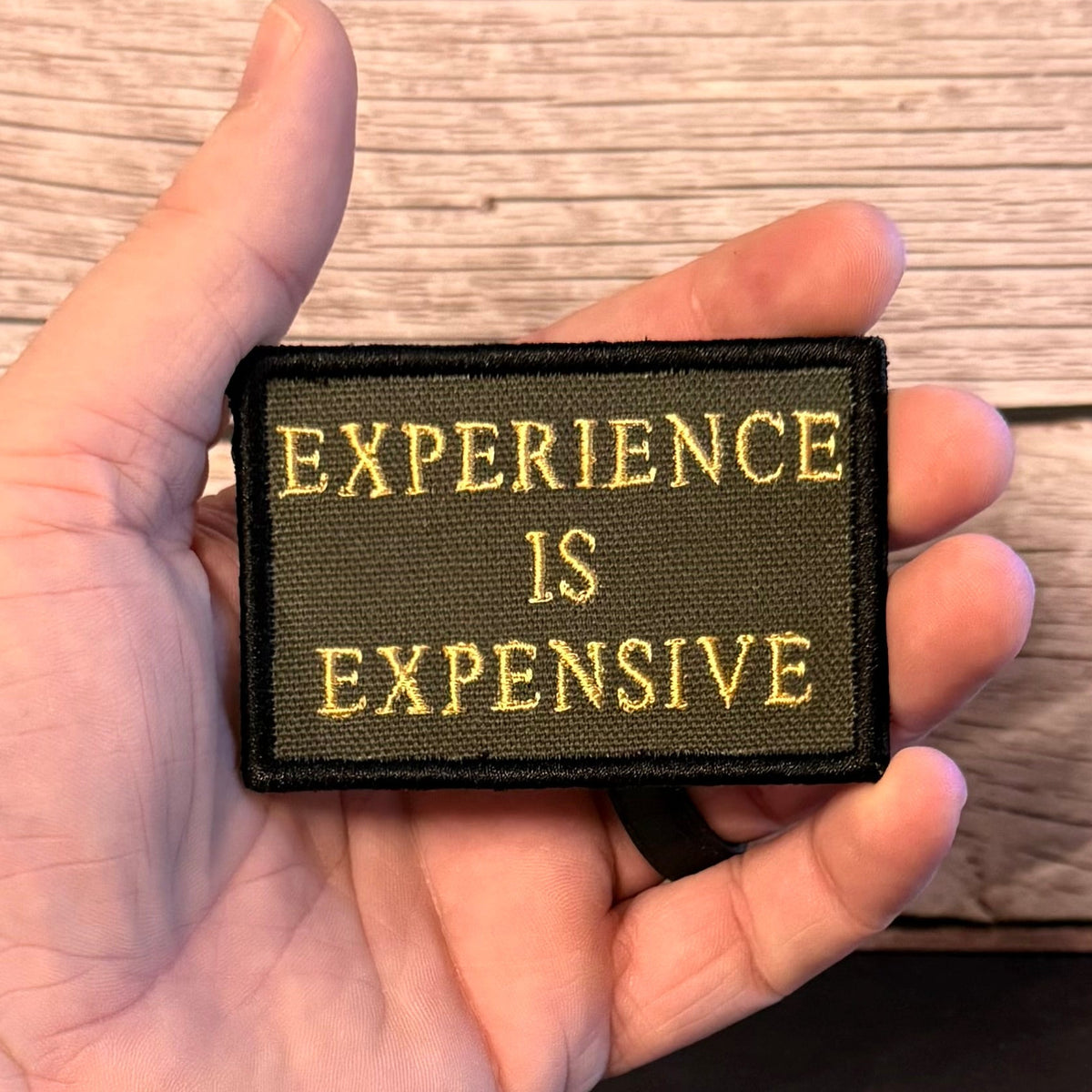 A hand holding a rectangular embroidered patch with the text 'EXPERIENCE IS EXPENSIVE' in gold lettering on a dark olive green background, set against a light wood plank background.