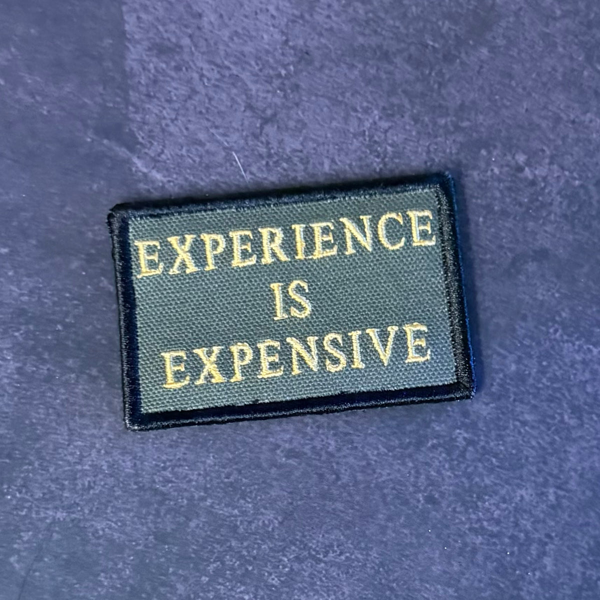 A rectangular embroidered patch with the text 'EXPERIENCE IS EXPENSIVE' in gold lettering on a dark olive green background, finished with a black stitched border, placed on a textured dark blue surface.