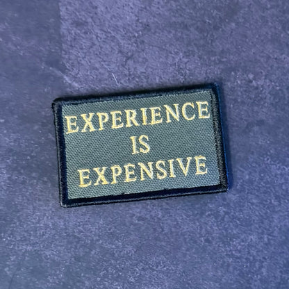 A rectangular embroidered patch with the text 'EXPERIENCE IS EXPENSIVE' in gold lettering on a dark olive green background, finished with a black stitched border, placed on a textured dark blue surface.