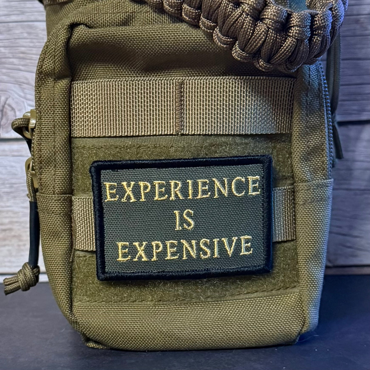 An olive green tactical pouch with a black rectangular patch that reads 'EXPERIENCE IS EXPENSIVE' in gold lettering. A brown paracord strap is threaded through the top zipper, and the pouch is photographed against a dark wood background.