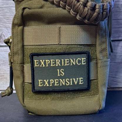 An olive green tactical pouch with a black rectangular patch that reads 'EXPERIENCE IS EXPENSIVE' in gold lettering. A brown paracord strap is threaded through the top zipper, and the pouch is photographed against a dark wood background.