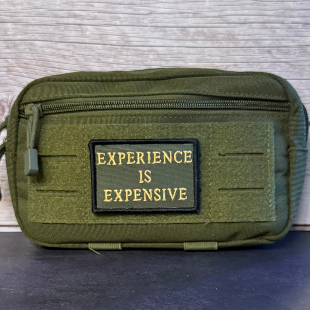 An olive green tactical pouch with a front zippered pocket and a rectangular patch reading 'EXPERIENCE IS EXPENSIVE' in yellow text, set against a light wood background.