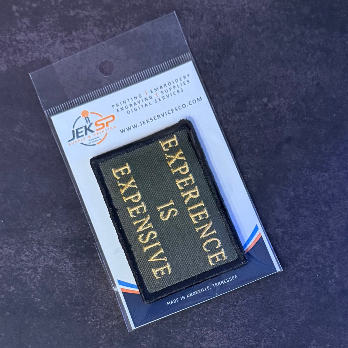 A rectangular embroidered patch with the words 'EXPERIENCE IS EXPENSIVE' in gold lettering on a black background, sealed in a clear plastic package with a branded card insert. The card shows the brand JEK SP.
