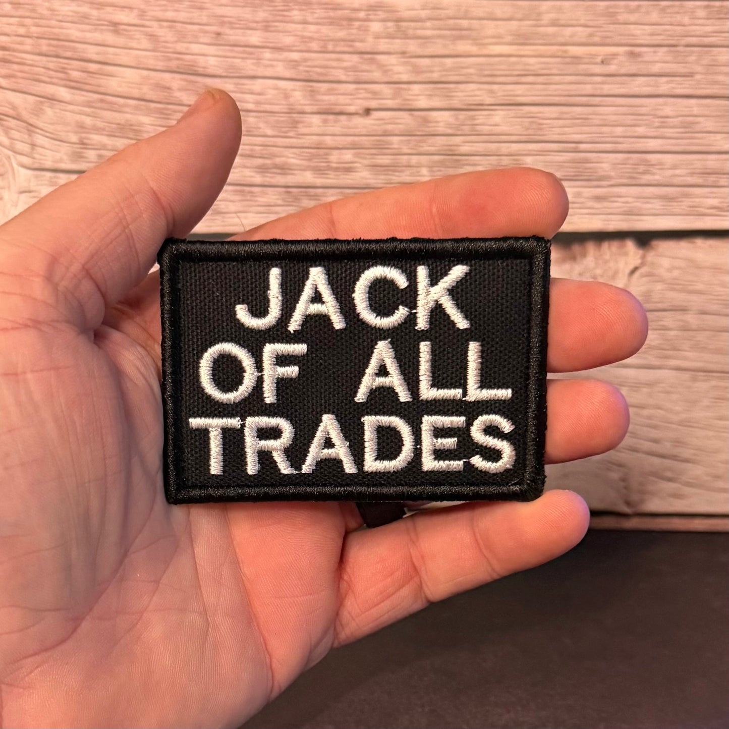 A hand holding a rectangular black embroidered patch with white text that reads 'JACK OF ALL TRADES' against a light wood background.