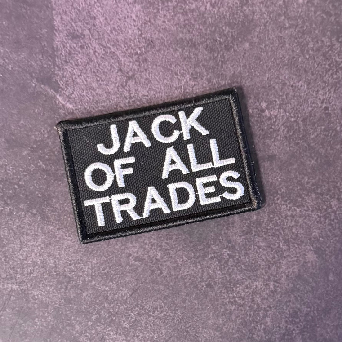 A rectangular black embroidered patch with the words 'JACK OF ALL TRADES' in white capital letters, placed on a textured purple surface.