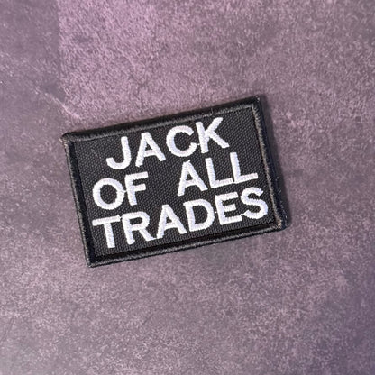 A rectangular black embroidered patch with the words 'JACK OF ALL TRADES' in white capital letters, placed on a textured purple surface.