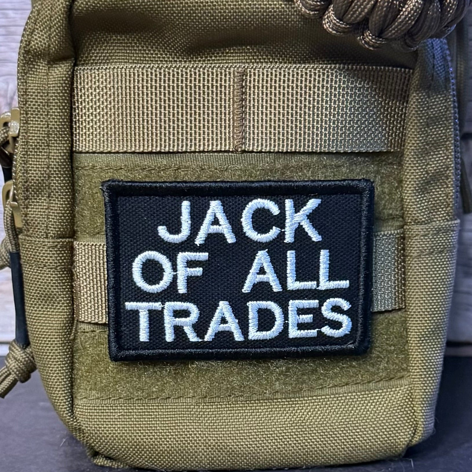 An olive green tactical pouch with a black embroidered patch reading 'JACK OF ALL TRADES' in white lettering, shown close-up against a rustic wood background.