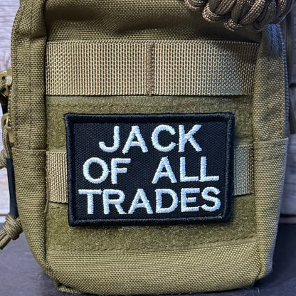 An olive green tactical pouch with a black embroidered patch reading 'JACK OF ALL TRADES' in white lettering, shown close-up against a rustic wood background.