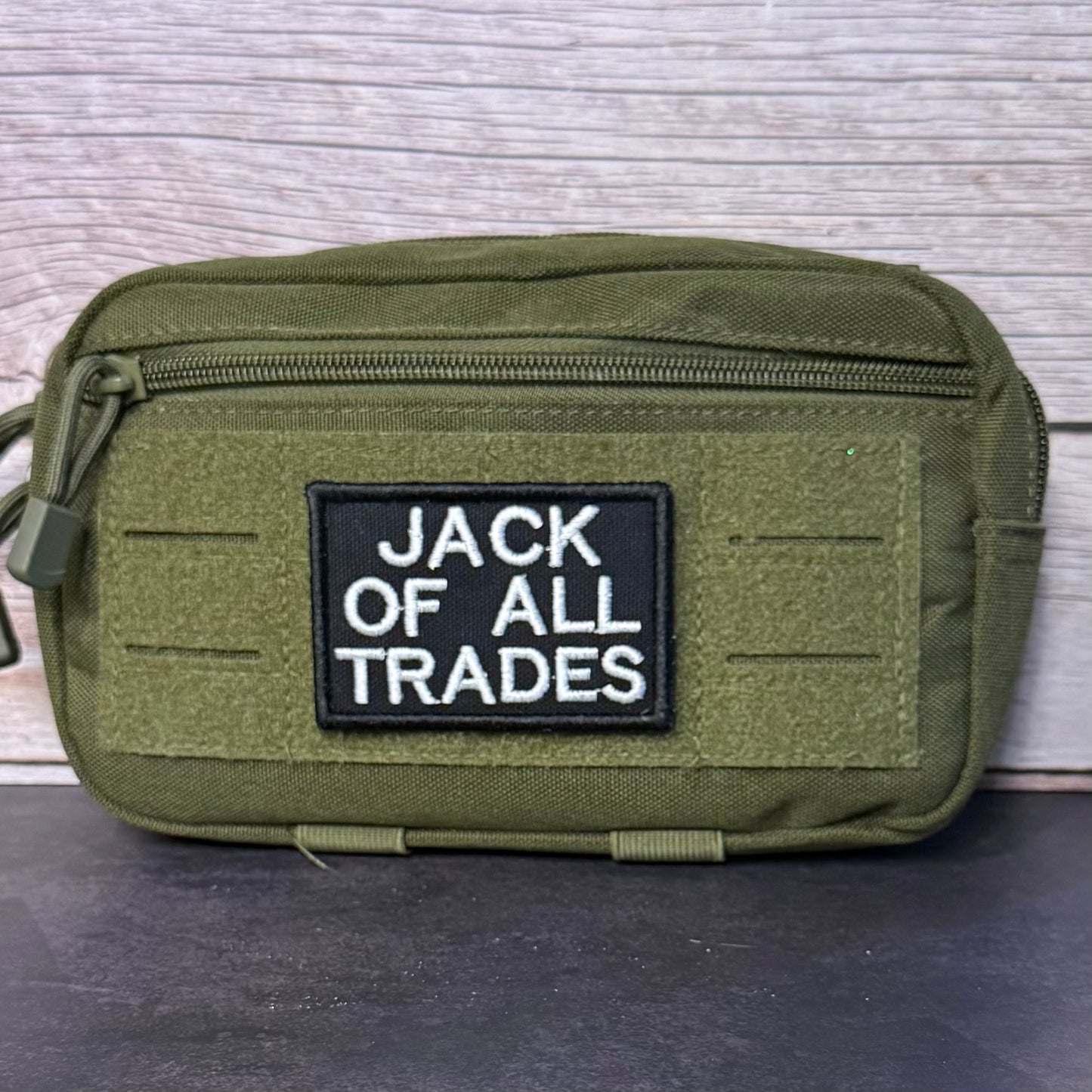 An olive green tactical pouch with a black rectangular patch reading 'JACK OF ALL TRADES' in white text, photographed against a light wood plank background.