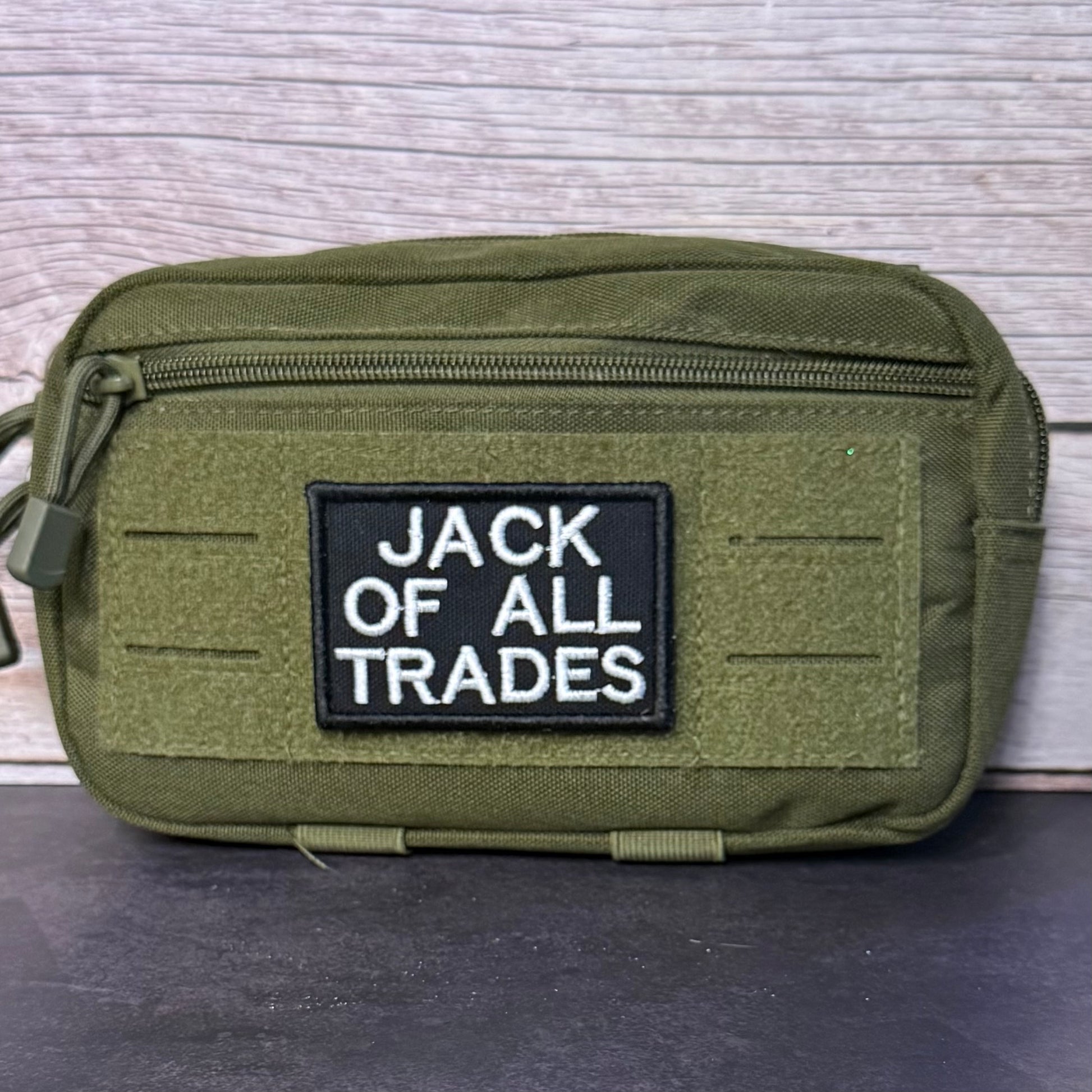An olive green tactical pouch with a black rectangular patch reading 'JACK OF ALL TRADES' in white text, photographed against a light wood plank background.