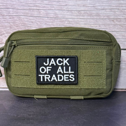 An olive green tactical pouch with a black rectangular patch reading 'JACK OF ALL TRADES' in white text, photographed against a light wood plank background.