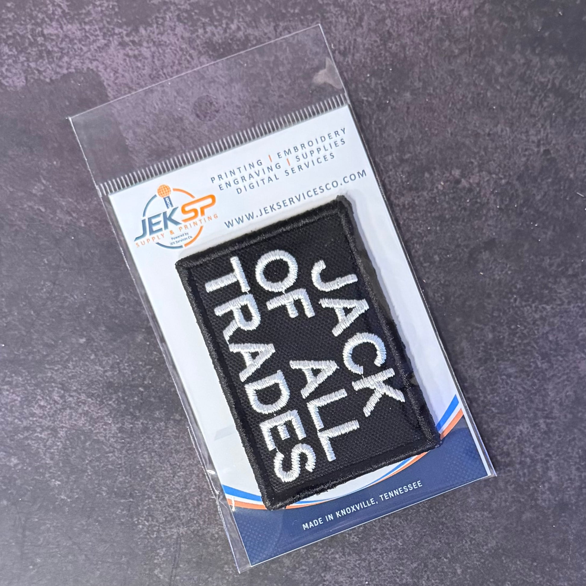 A black rectangular embroidered patch with white text reading 'JACK OF ALL TRADES' inside a clear plastic sleeve on a branded card, photographed on a dark textured surface.