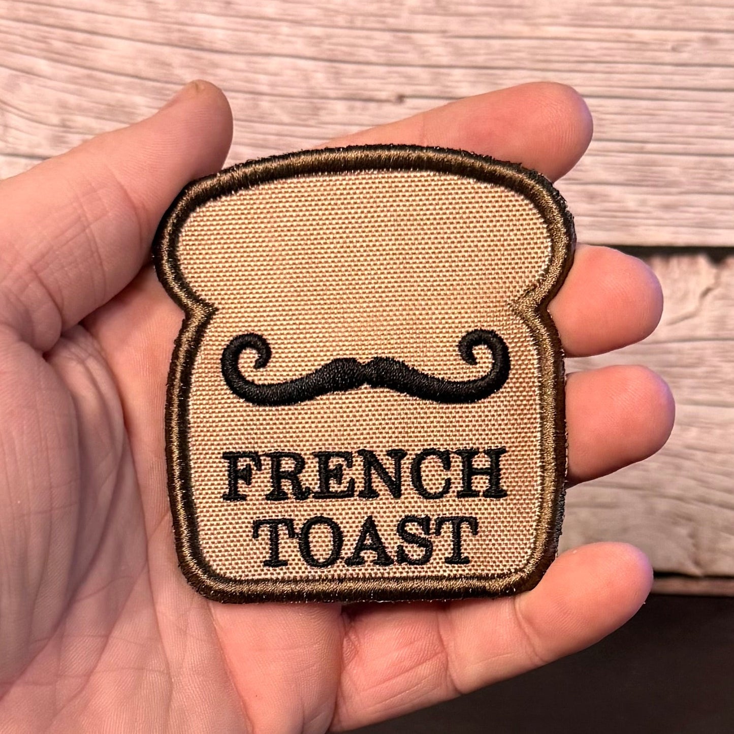 A hand holding a small embroidered patch shaped like a slice of French toast with a black mustache and the words 'FRENCH TOAST' on a tan woven fabric background, against a wood plank backdrop.