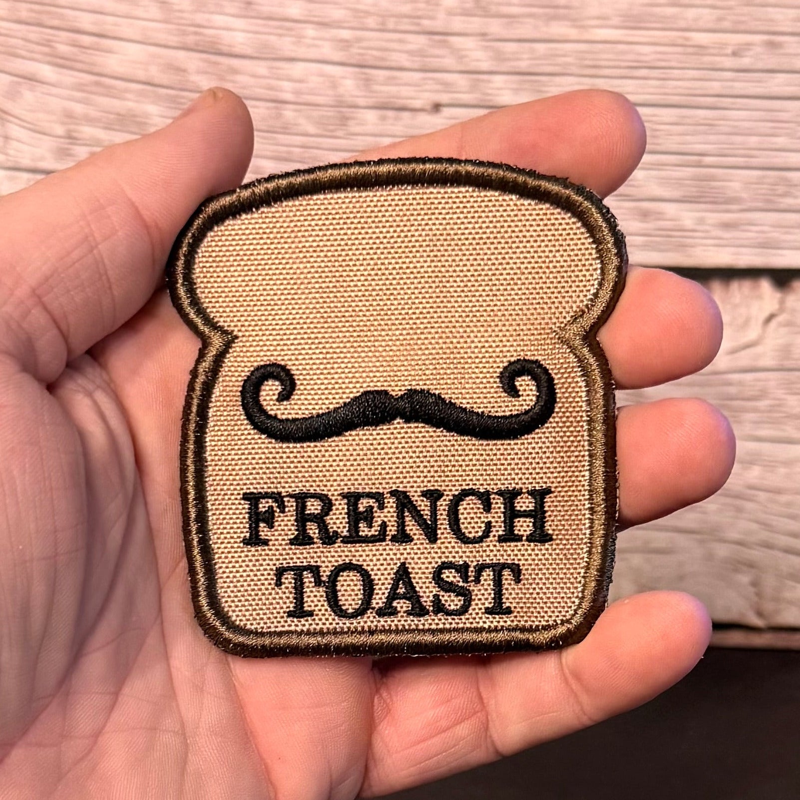 A hand holding a small embroidered patch shaped like a slice of French toast with a black mustache and the words 'FRENCH TOAST' on a tan woven fabric background, against a wood plank backdrop.
