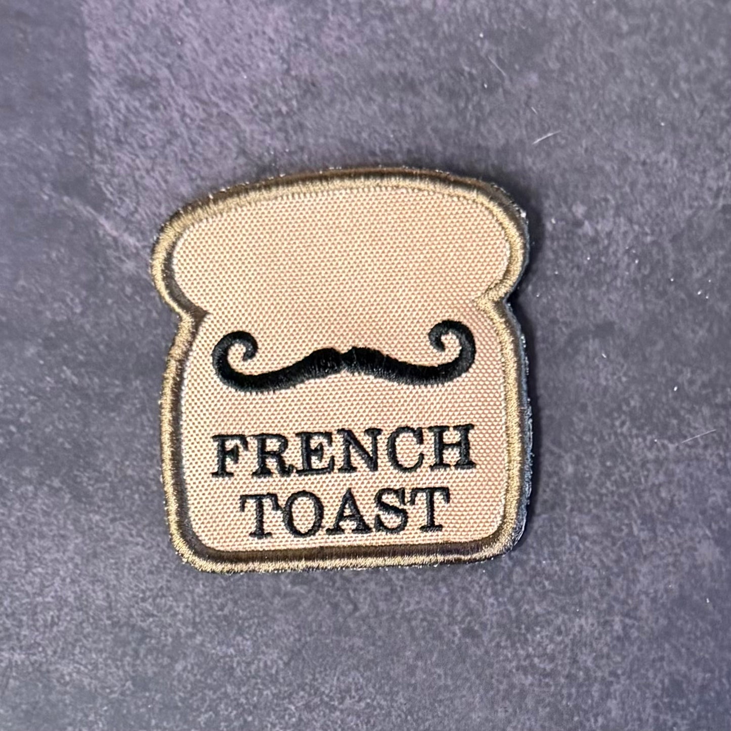 A bread-slice-shaped embroidered patch with a black mustache and the words 'FRENCH TOAST' on a dark textured background.