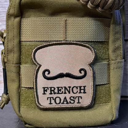 An olive green tactical pouch with a woven paracord strap, featuring a tan embroidered patch shaped like a French toast slice with a black mustache and the text 'FRENCH TOAST'.