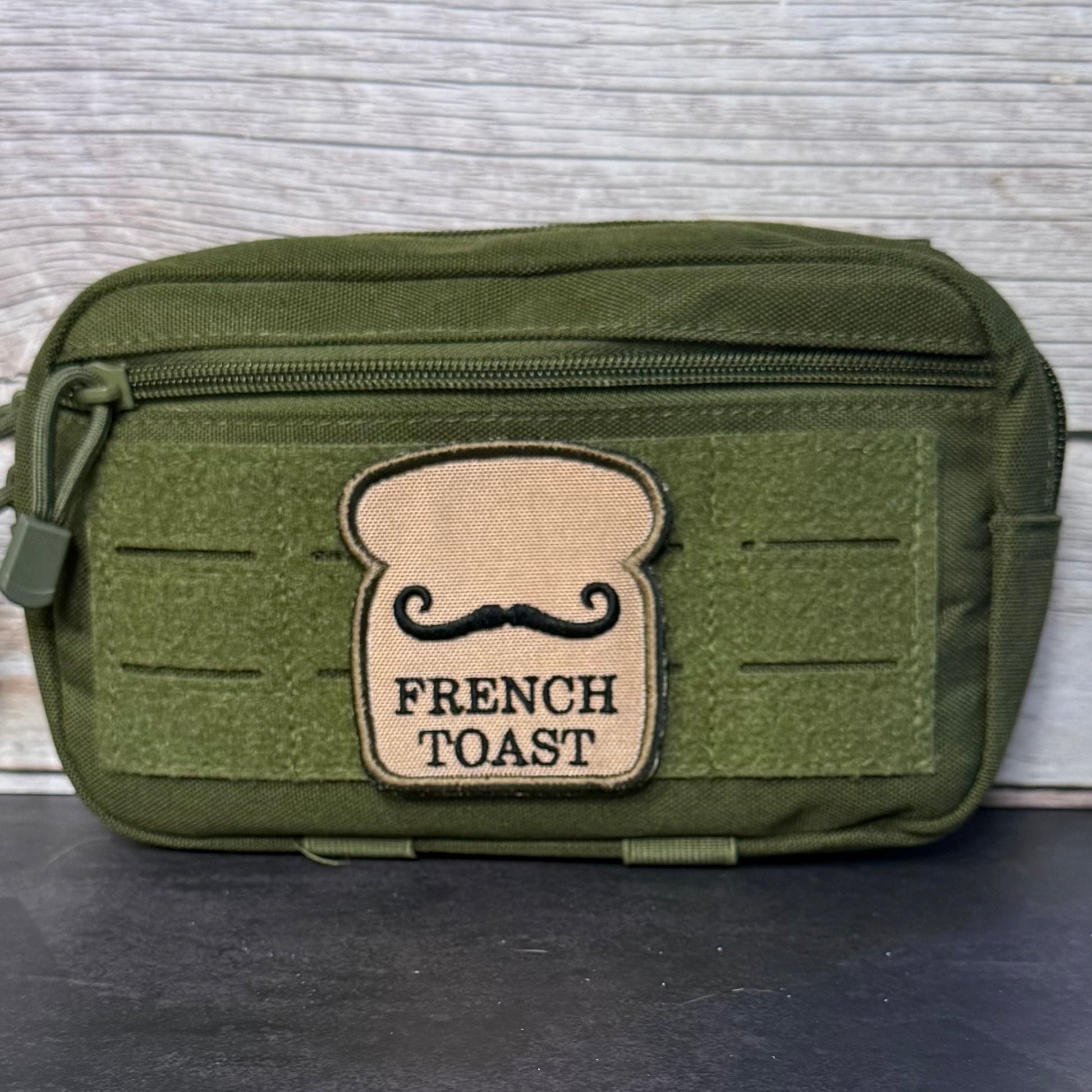 An olive green tactical pouch with a top zipper and a tan patch shaped like a slice of bread with a black mustache and the words 'FRENCH TOAST' on the front, set against a light gray wood background.