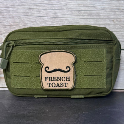 An olive green tactical pouch with a top zipper and a tan patch shaped like a slice of bread with a black mustache and the words 'FRENCH TOAST' on the front, set against a light gray wood background.