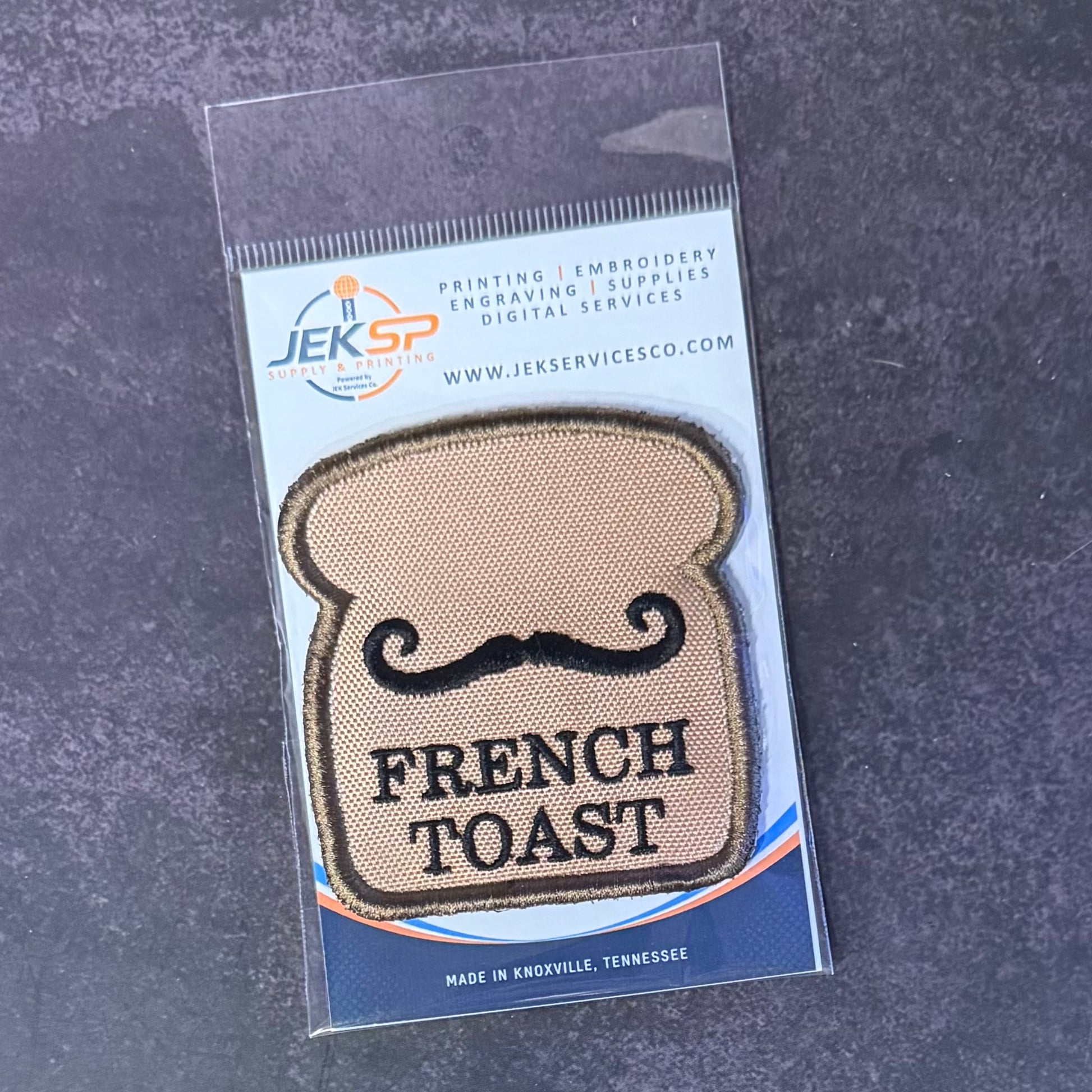 An embroidered patch shaped like a slice of French toast with a black mustache and the words 'FRENCH TOAST' in black, sealed in a clear plastic package with a blue and white Jeksp Service Co. backing card, placed on a dark textured surface.