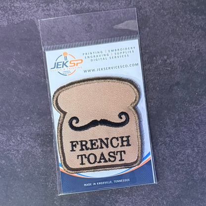 An embroidered patch shaped like a slice of French toast with a black mustache and the words 'FRENCH TOAST' in black, sealed in a clear plastic package with a blue and white Jeksp Service Co. backing card, placed on a dark textured surface.