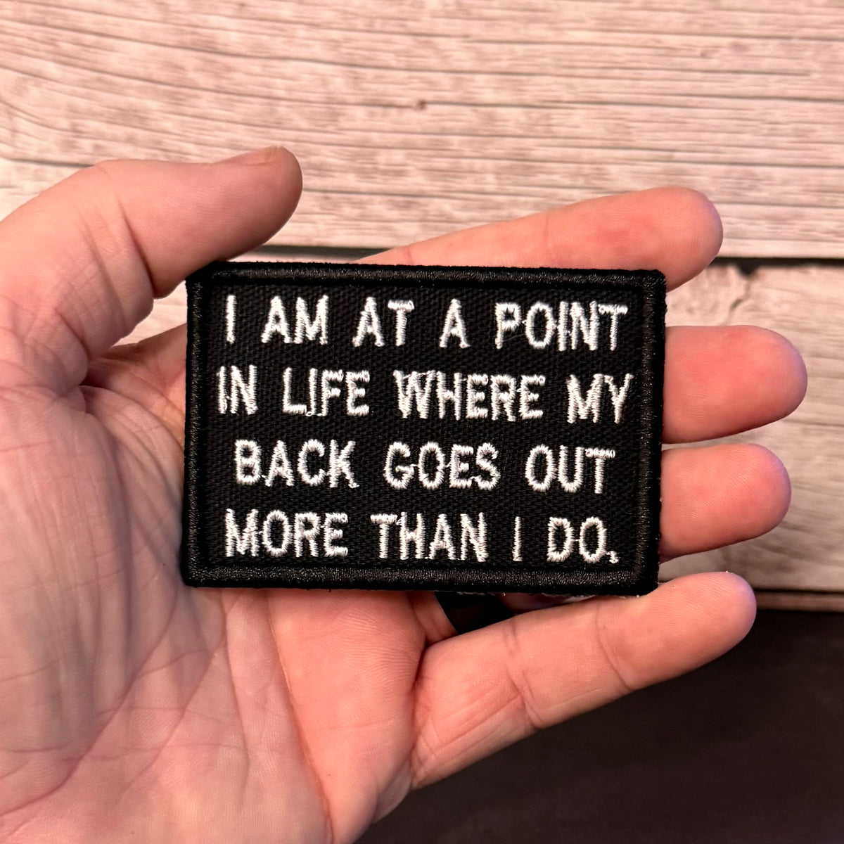 A hand holding a rectangular black embroidered patch with white text that reads, 'I AM AT A POINT IN LIFE WHERE MY BACK GOES OUT MORE THAN I DO.' The background shows a light wood surface.