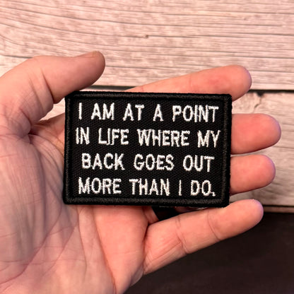 A hand holding a rectangular black embroidered patch with white text that reads, 'I AM AT A POINT IN LIFE WHERE MY BACK GOES OUT MORE THAN I DO.' The background shows a light wood surface.