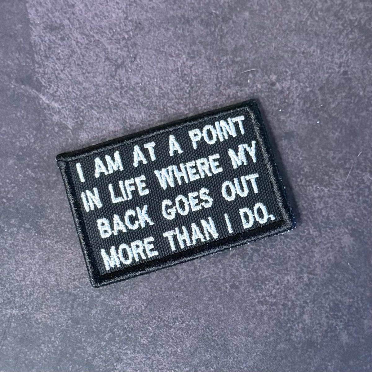 A rectangular black embroidered patch with white text reading 'I AM AT A POINT IN LIFE WHERE MY BACK GOES OUT MORE THAN I DO.' placed on a textured gray surface.