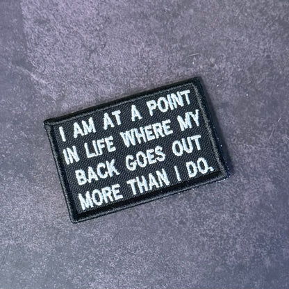 A rectangular black embroidered patch with white text reading 'I AM AT A POINT IN LIFE WHERE MY BACK GOES OUT MORE THAN I DO.' placed on a textured gray surface.