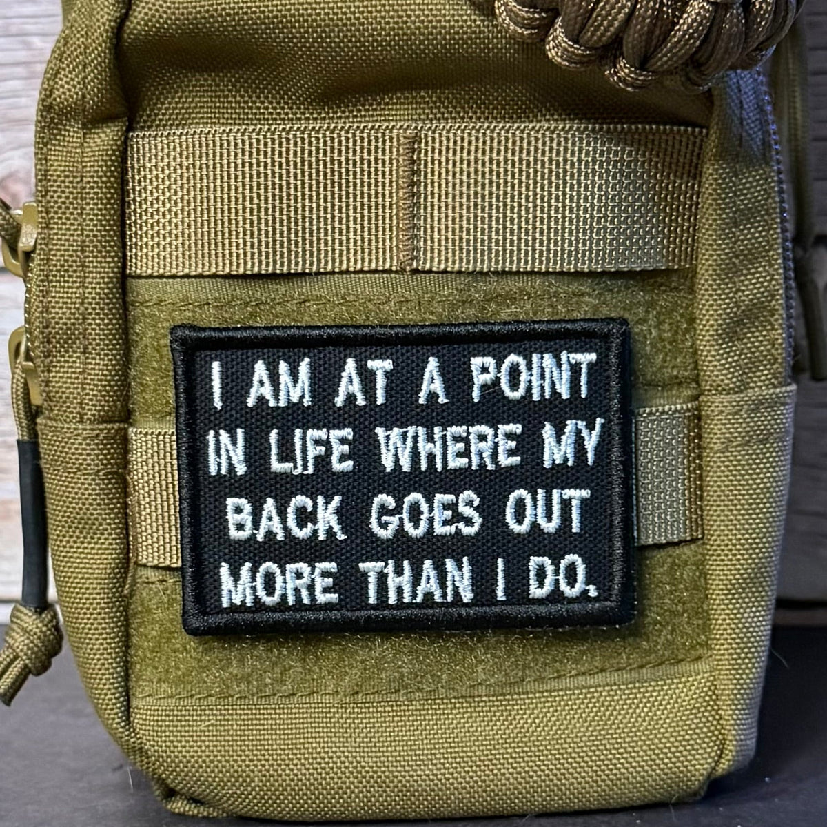 An olive drab tactical pouch with a black rectangular patch featuring white embroidered text that reads, 'I AM AT A POINT IN LIFE WHERE MY BACK GOES OUT MORE THAN I DO.' A braided tan strap is attached at the top, and the pouch is photographed against a rustic wood background.