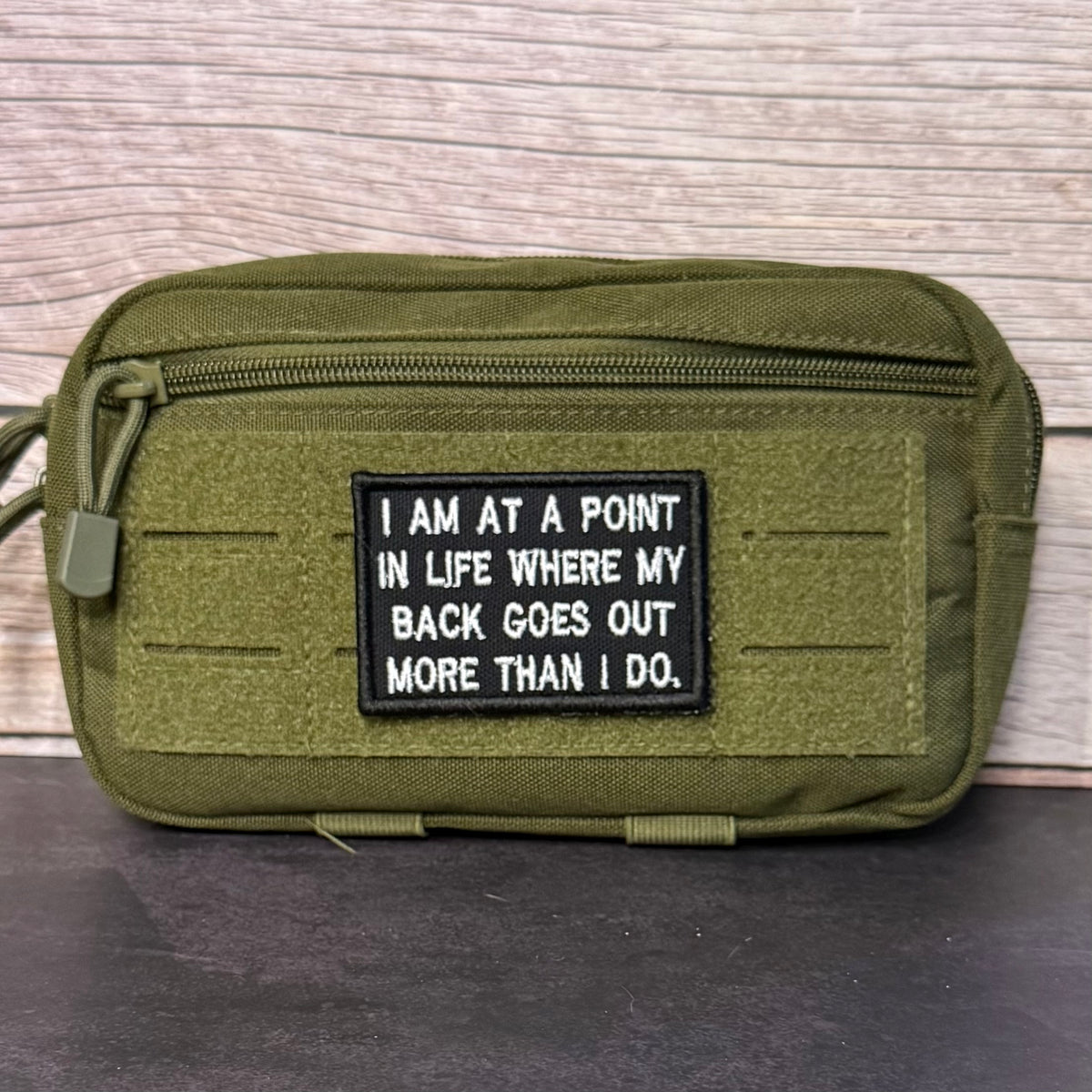 An olive green tactical pouch with a black rectangular patch featuring white text that reads, 'I AM AT A POINT IN LIFE WHERE MY BACK GOES OUT MORE THAN I DO.' The pouch is shown against a light wood panel background.