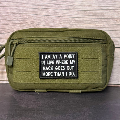 An olive green tactical pouch with a black rectangular patch featuring white text that reads, 'I AM AT A POINT IN LIFE WHERE MY BACK GOES OUT MORE THAN I DO.' The pouch is shown against a light wood panel background.