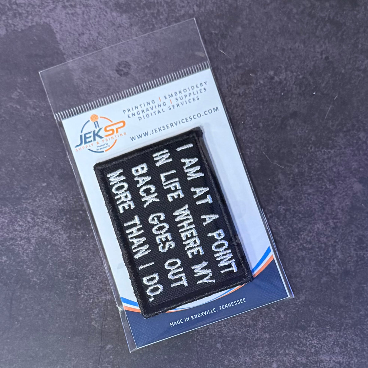 A black rectangular embroidered patch with white text reading 'I AM AT A POINT IN LIFE WHERE MY BACK GOES OUT MORE THAN I DO.' displayed inside a clear plastic retail sleeve on a branded card, photographed on a dark textured surface.