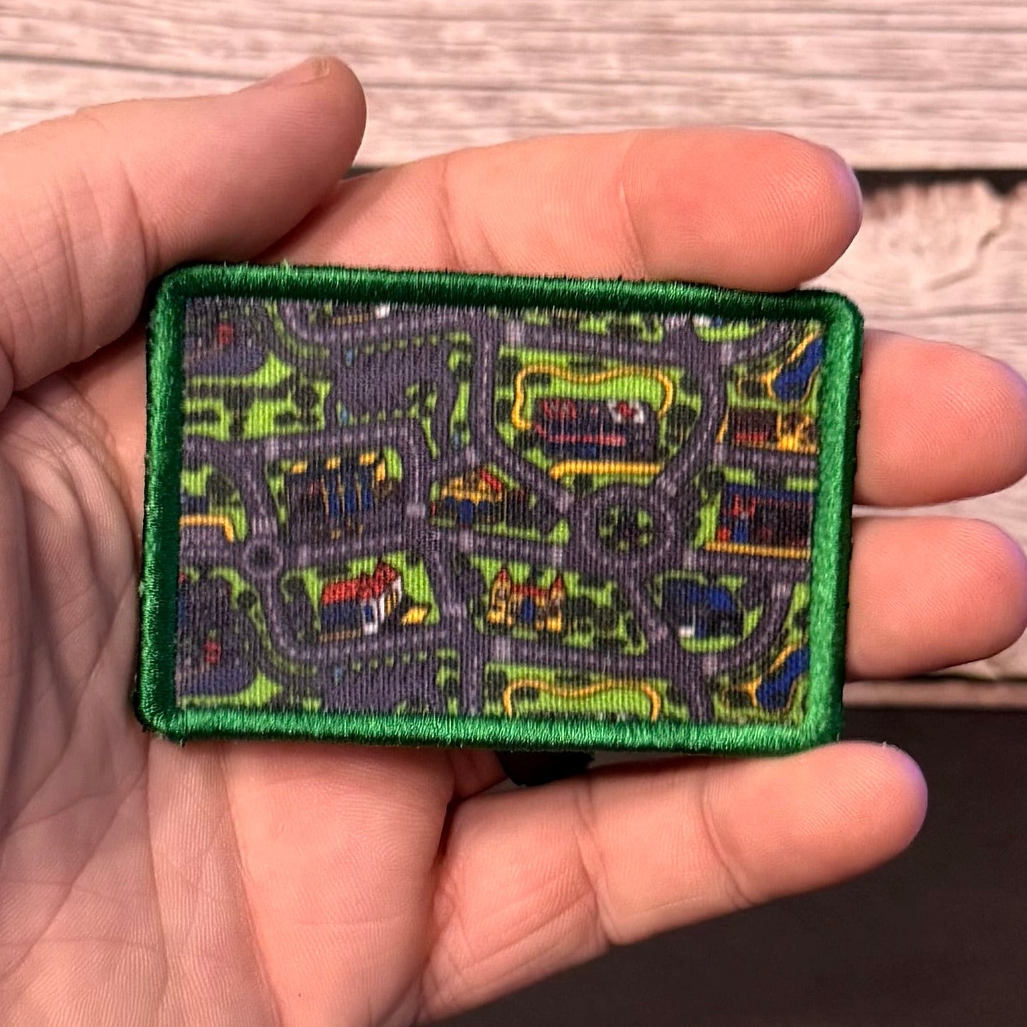A hand holding a rectangular embroidered patch with a green border and a detailed map design in gray, green, yellow, and red, set against a light wood background.