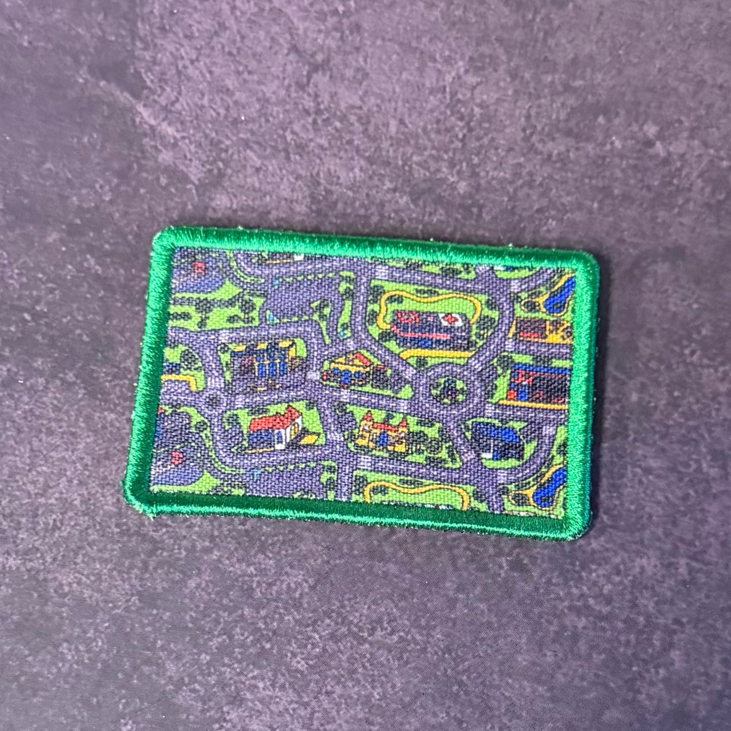 A rectangular embroidered patch with a green border and a multicolor map-like design featuring roads, buildings, and green areas, placed on a textured gray surface.