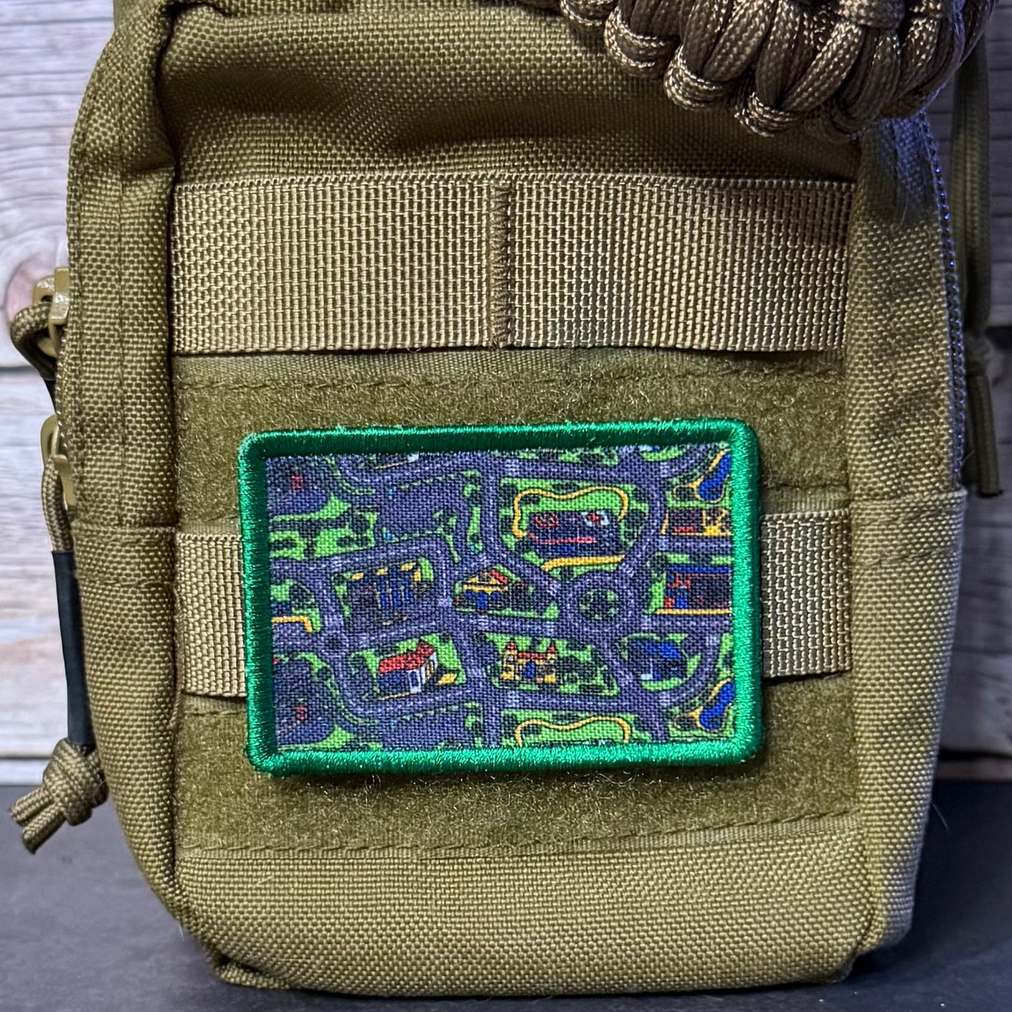 An embroidered tactical patch featuring a colorful map design with a bright green border, attached to the front of an olive drab nylon pouch with webbing.