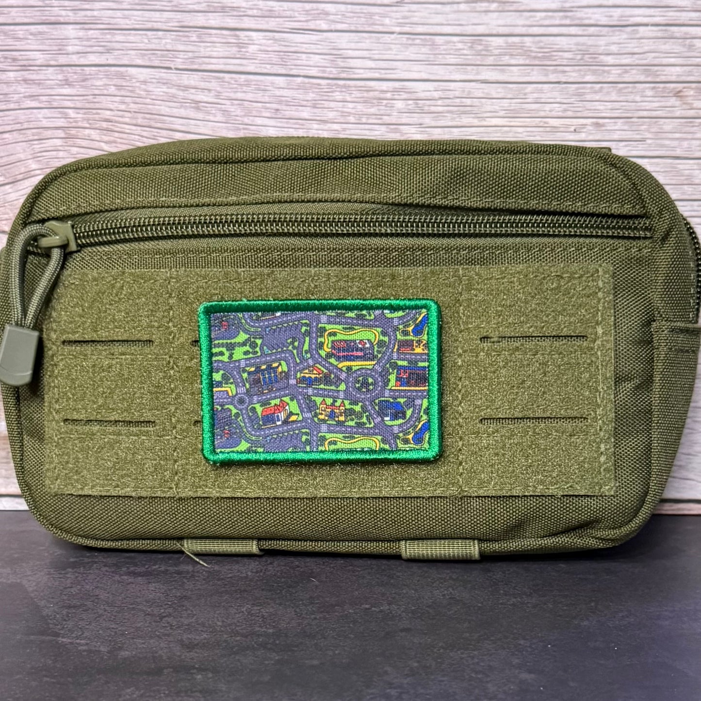 An olive green tactical pouch with a zippered top and a colorful embroidered patch featuring a map design, set against a light wood background.