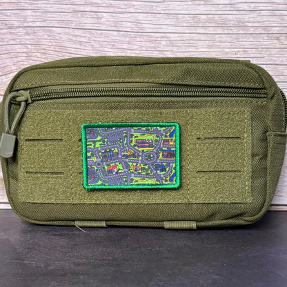 An olive green tactical pouch with a zippered top and a colorful embroidered patch featuring a map design, set against a light wood background.