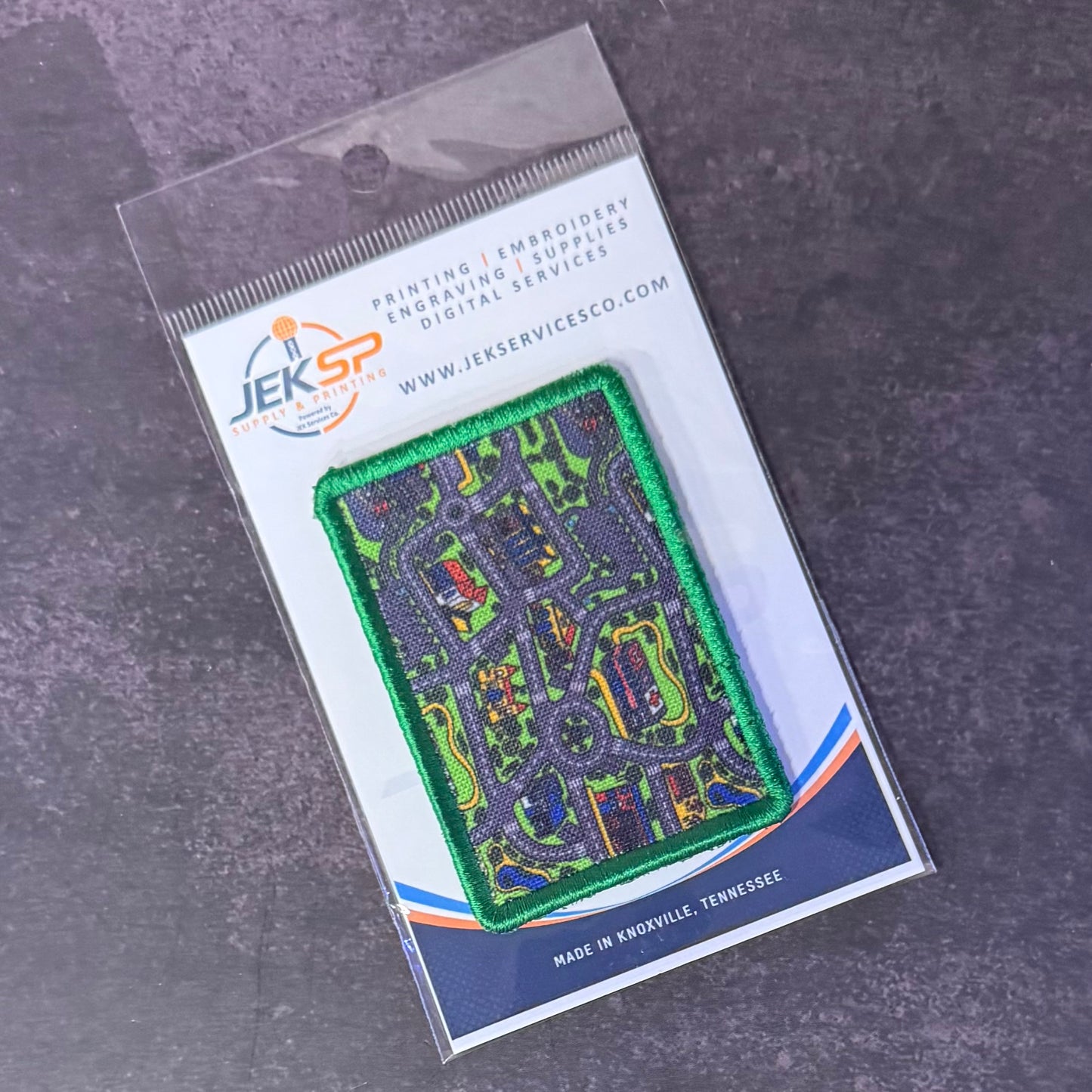 An embroidered rectangular patch showing a street map design in green, blue, and orange, packaged on a branded clear plastic card from JEKSP Embroidery Services against a dark gray textured background.