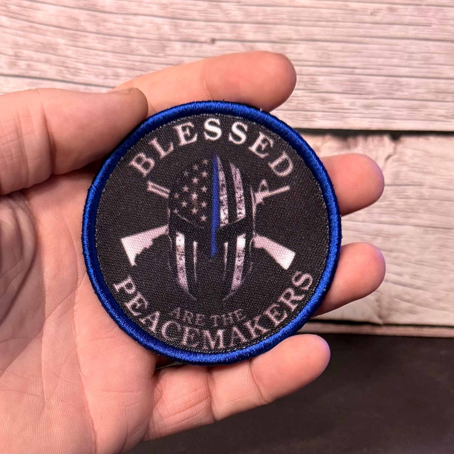A hand holding a circular embroidered patch with a blue border and a black background. The patch features a distressed American flag design with a single blue stripe down the center, crossed rifles, and the text 'BLESSED PEACEMAKERS ARE THE'.