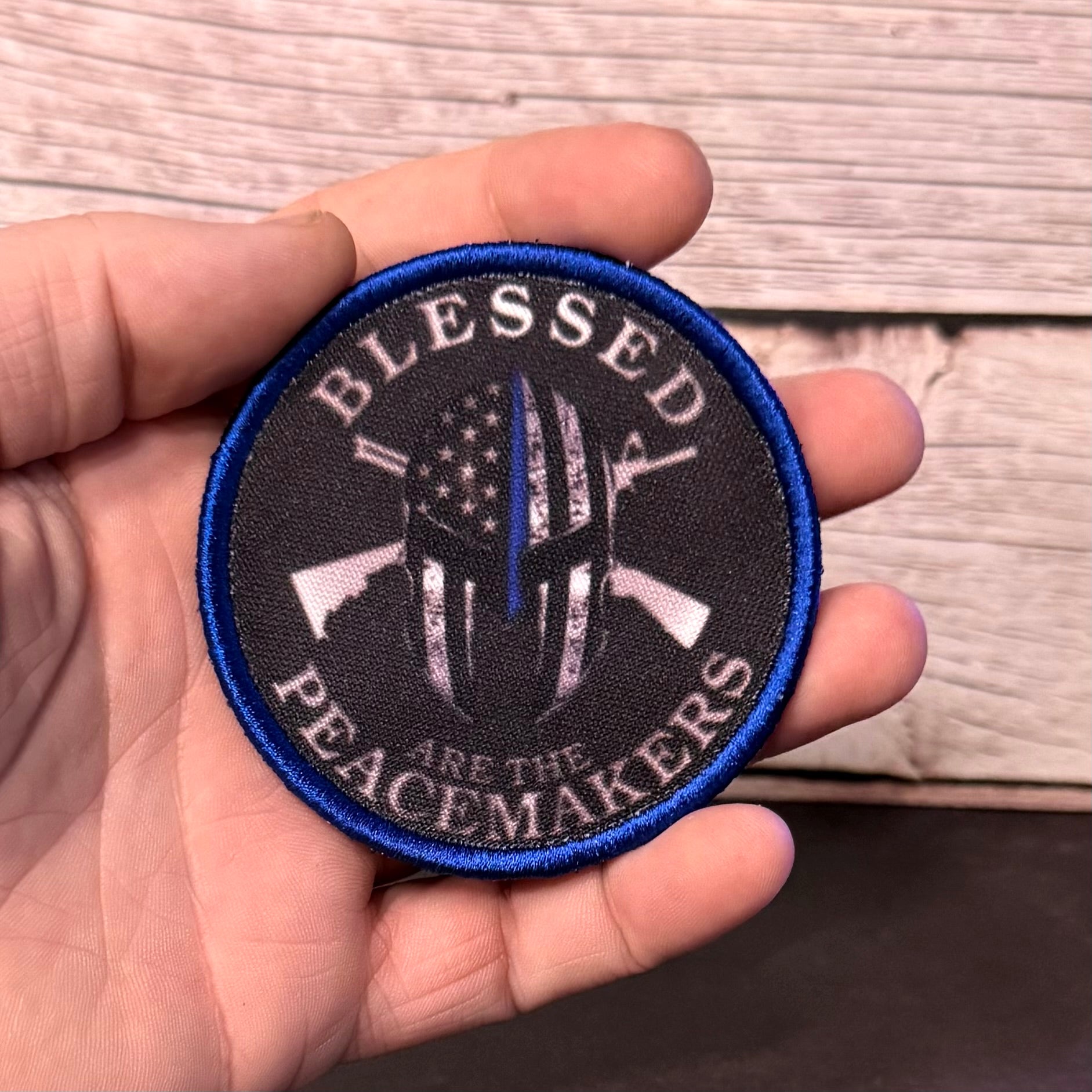 A hand holding a circular embroidered patch with a blue border and a black background. The patch features a distressed American flag design with a single blue stripe down the center, crossed rifles, and the text 'BLESSED PEACEMAKERS ARE THE'.