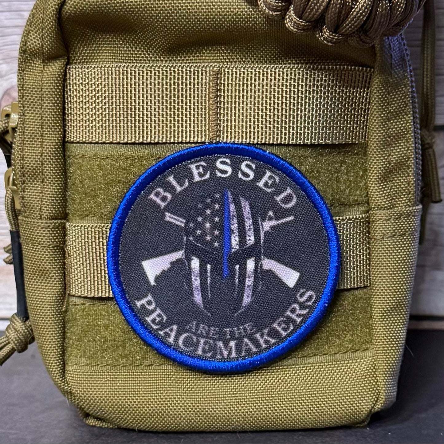 An olive green tactical pouch with a circular embroidered patch that reads 'BLESSED PEACEMAKERS' around a black-and-white helmet design with a blue outline, shown against a rustic wood background.