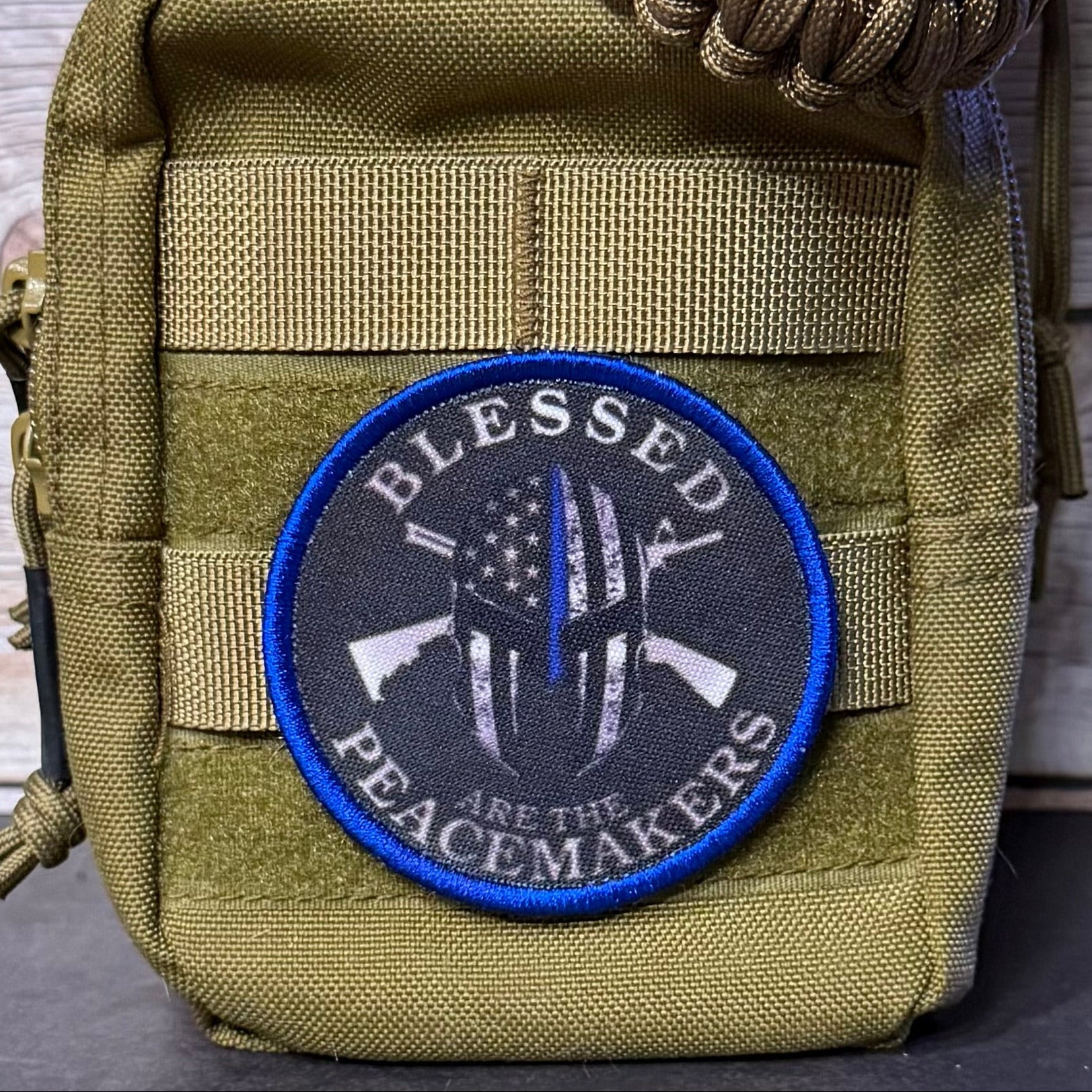 An olive green tactical pouch with a circular embroidered patch that reads 'BLESSED PEACEMAKERS' around a black-and-white helmet design with a blue outline, shown against a rustic wood background.