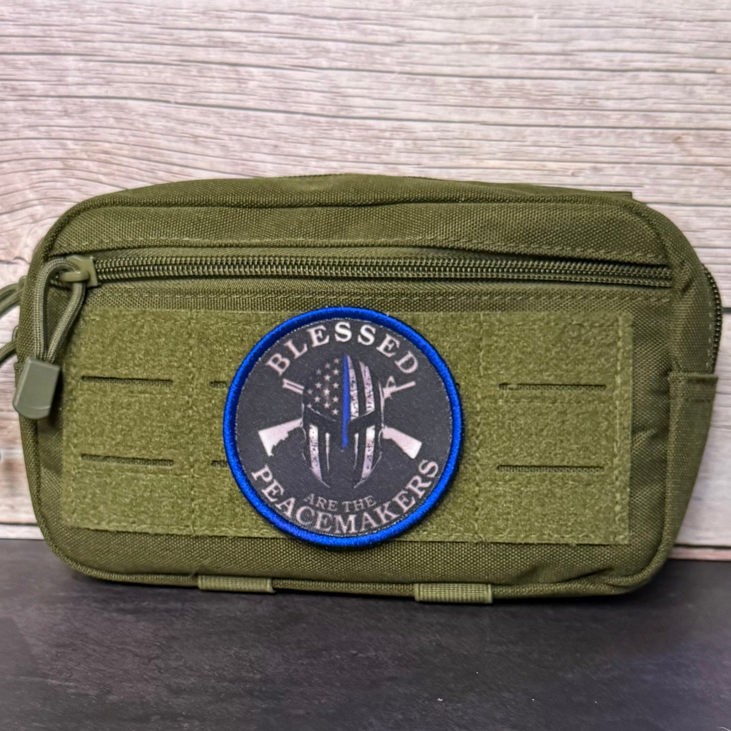An olive green tactical pouch with a front zipper and a circular embroidered patch that reads 'BLESSED PEACEMakers' with crossed rifles and a flag design, set against a light wood background.