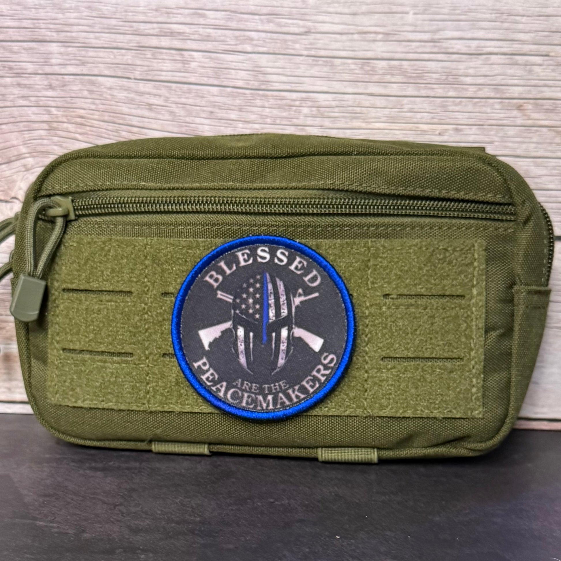 An olive green tactical pouch with a front zipper and a circular embroidered patch that reads 'BLESSED PEACEMakers' with crossed rifles and a flag design, set against a light wood background.