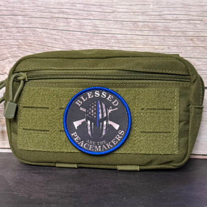 An olive green tactical pouch with a front zipper and a circular embroidered patch that reads 'BLESSED PEACEMakers' with crossed rifles and a flag design, set against a light wood background.